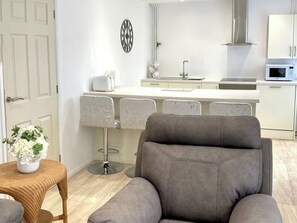 Cocina moderna con armarios blancos, microondas y campana extractora de acero inoxidable. Hay un comedor con una silla tapizada de color gris y una mesa de mimbre con una planta en maceta.