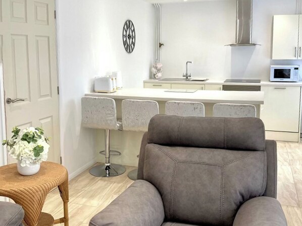 A modern kitchen with white cabinets, a microwave, and a stainless steel range hood. There is a dining area with a grey upholstered chair and a wicker table with a potted plant.