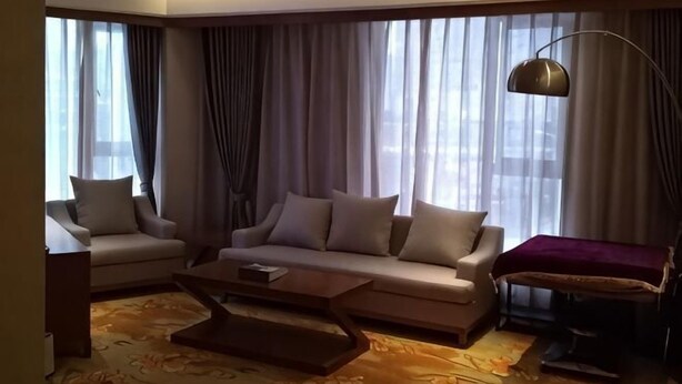 Una sala de estar moderna con sofá, sillón y mesa de centro, ventanas amplias con cortinas y una lámpara de pie.