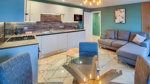 Eine moderne Küche mit weißen Schränken, einem Einbauofen und einem Spülbecken. Es gibt einen Essbereich mit einem Glastisch und Stühlen, sowie ein graues Sofa im Wohnzimmer.