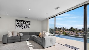 A modern living room with a grey sectional sofa, a large abstract artwork, and a view of a swimming pool and houses.