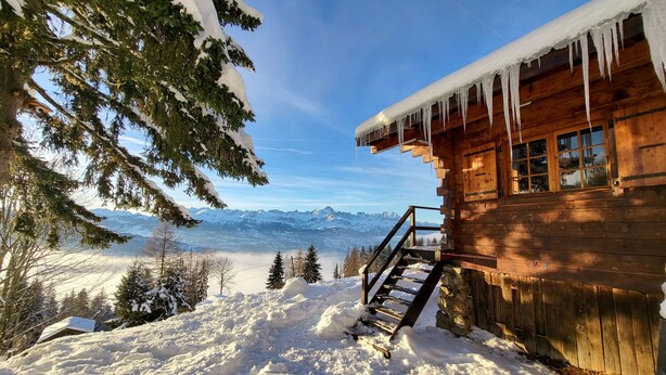 A wooden cabin with a staircase leading to a deck, surrounded by snow-covered trees and a mountain view.