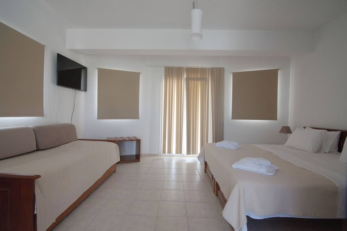 A hotel room with a sofa, a bed, a TV mounted on the wall, and a window with curtains.