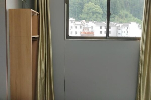 A room with a window, a wooden wardrobe, and a view of buildings and greenery outside.
