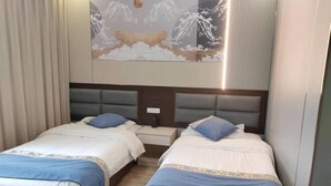 Hotellihuone, jossa on kaksi sänkyä, yöpöytä ja taulu, jossa on kuvattuna lumihuippuinen vuoristomaisema.