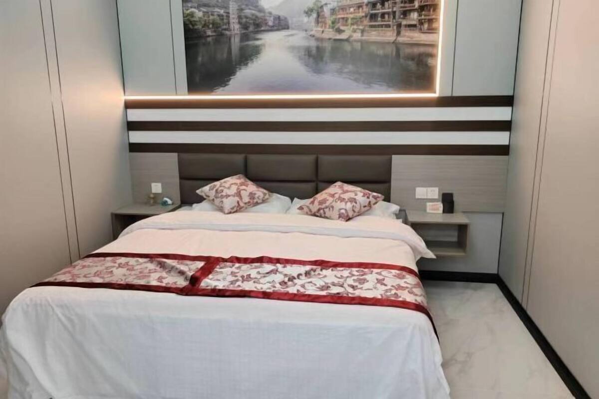 A hotel room with a bed, a nightstand, a shelf, and a large window with a view of a river and buildings.