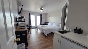 A hotel room with a bed, a desk, a kitchenette, and a ceiling fan.