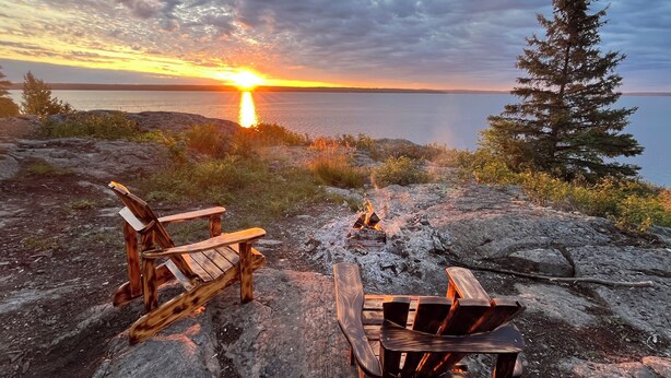 Two wooden Adirondack chairs on a rocky shore with a sunset in the background.