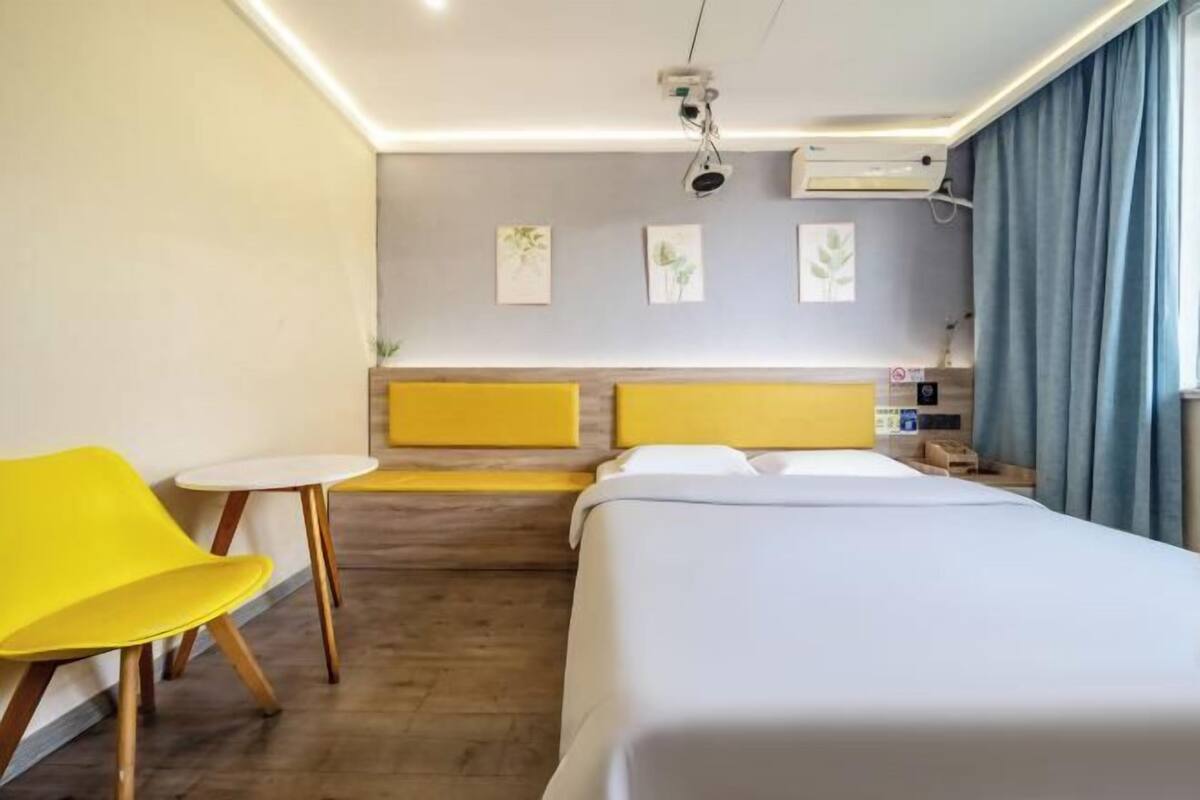 A hotel room with a bed, a small table, a yellow chair, and a headboard with a light fixture.