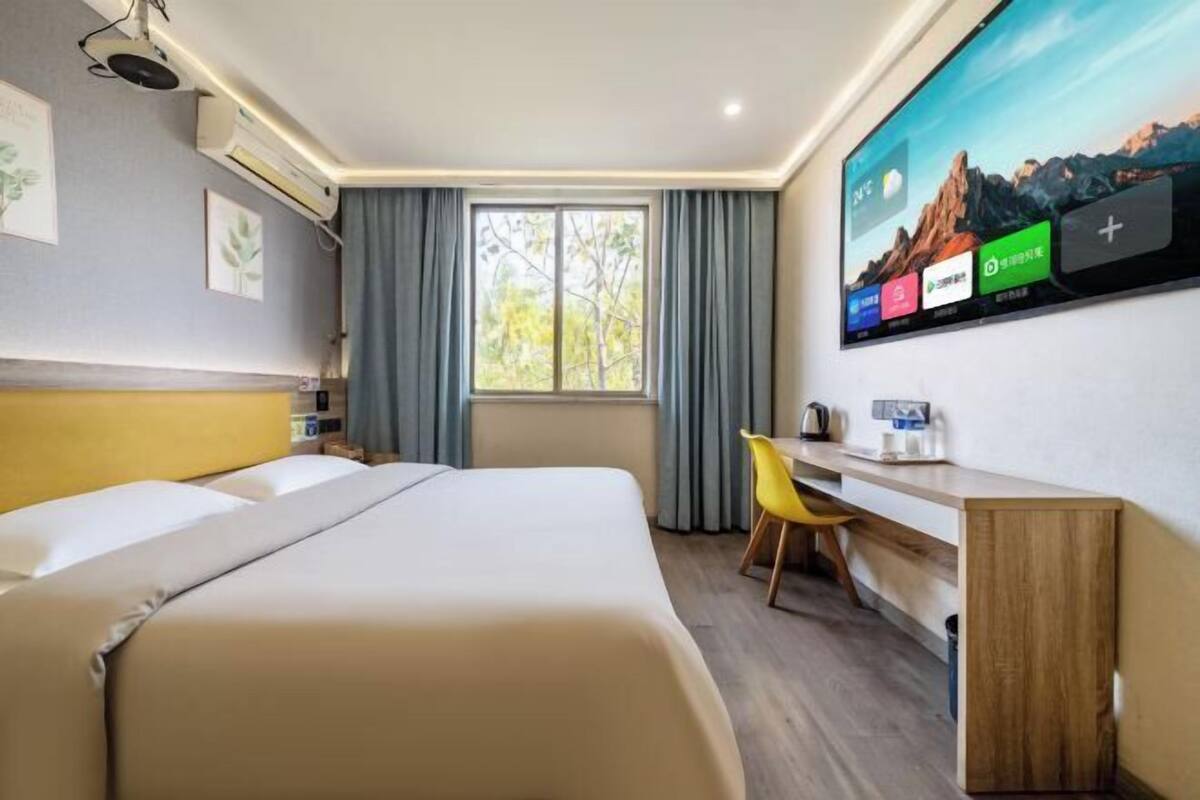 A hotel room with a bed, a desk, a chair, a TV mounted on the wall, and a window with curtains.
