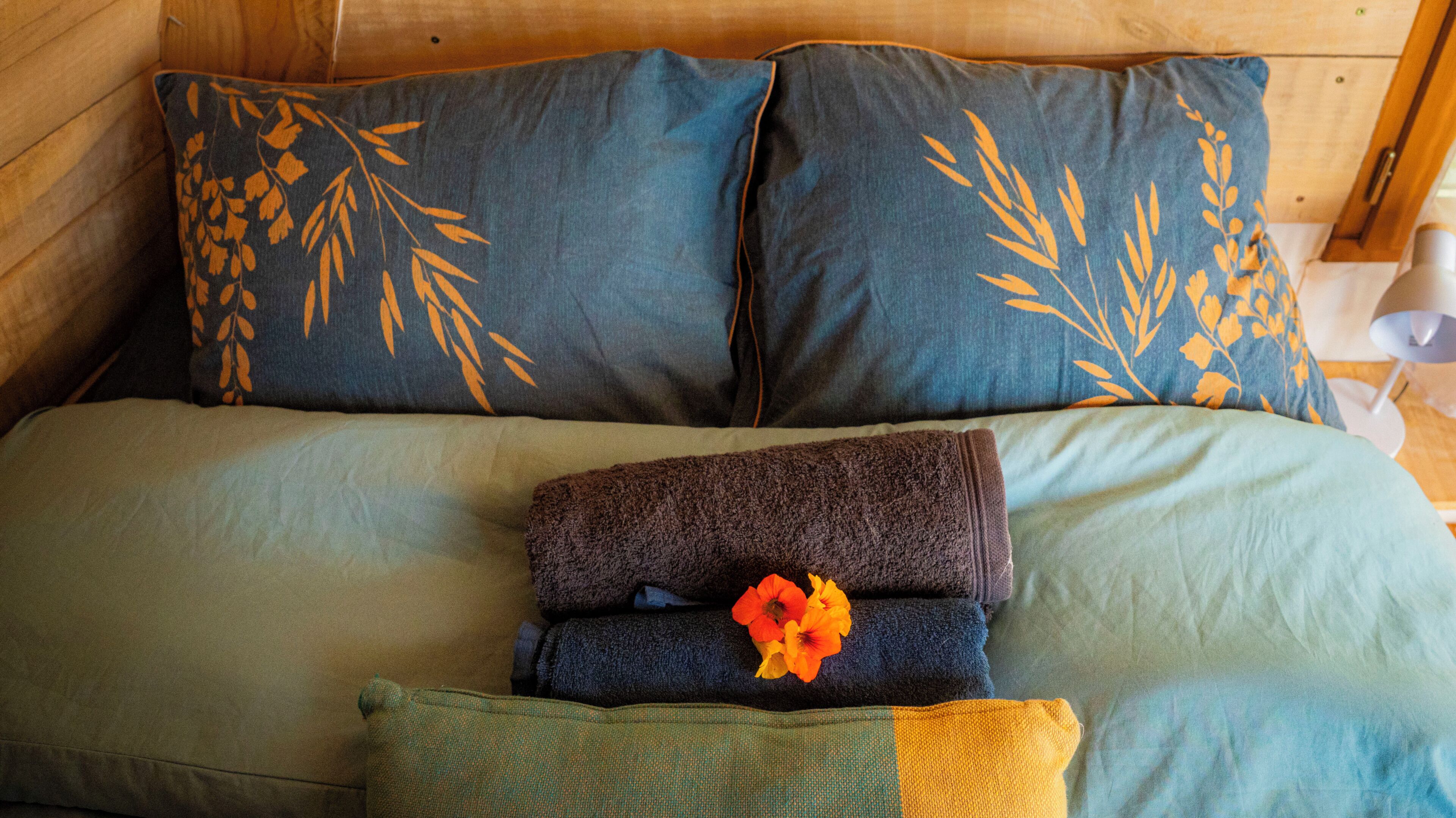 Una cama con una colcha azul y verde, dos almohadas con estampado de hojas y una toalla doblada con un motivo floral.