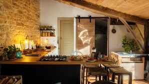 A rustic kitchen with a stone wall, wooden beams, a gas stove, and a wooden dining area with chairs.