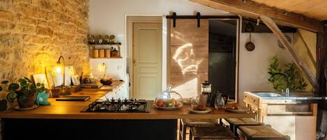 A rustic kitchen with a stone wall, wooden beams, a gas stove, and a wooden dining area with chairs.