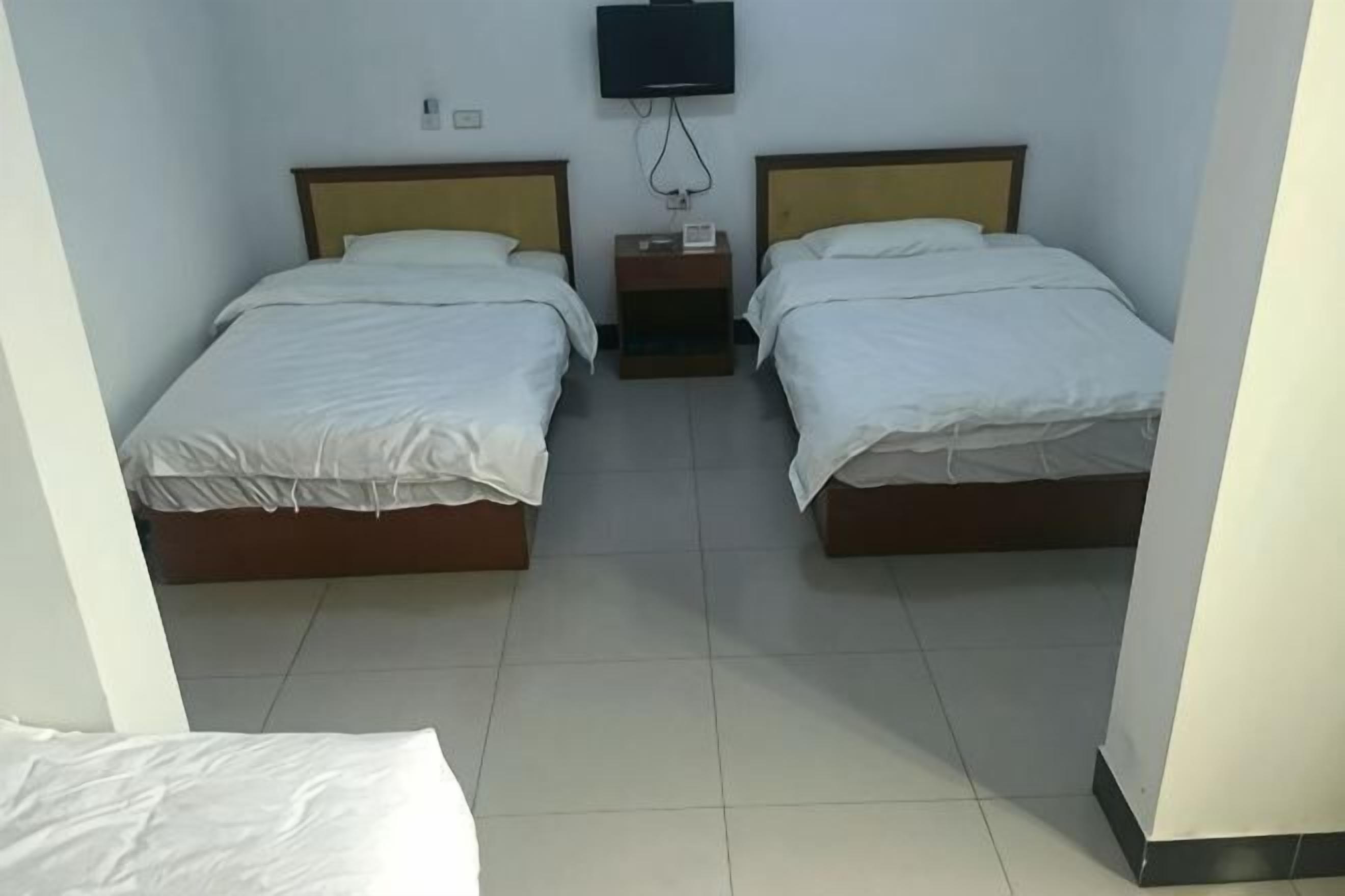 A room with two beds, a flat-screen TV mounted on the wall, and a small bedside table with a lamp.