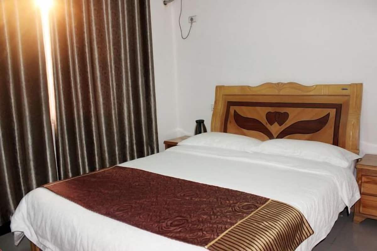 Habitación de hotel con cabecera de madera, cama con ropa de cama blanca y marrón, mesita de noche y cortina.