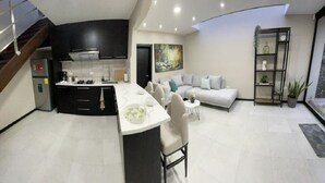 A modern kitchen with a central island, a dining area with a white table and chairs, a grey sofa, and a staircase on the left. - 2BR duplex with city views.  "Casa Colibrí" (Cuenca)