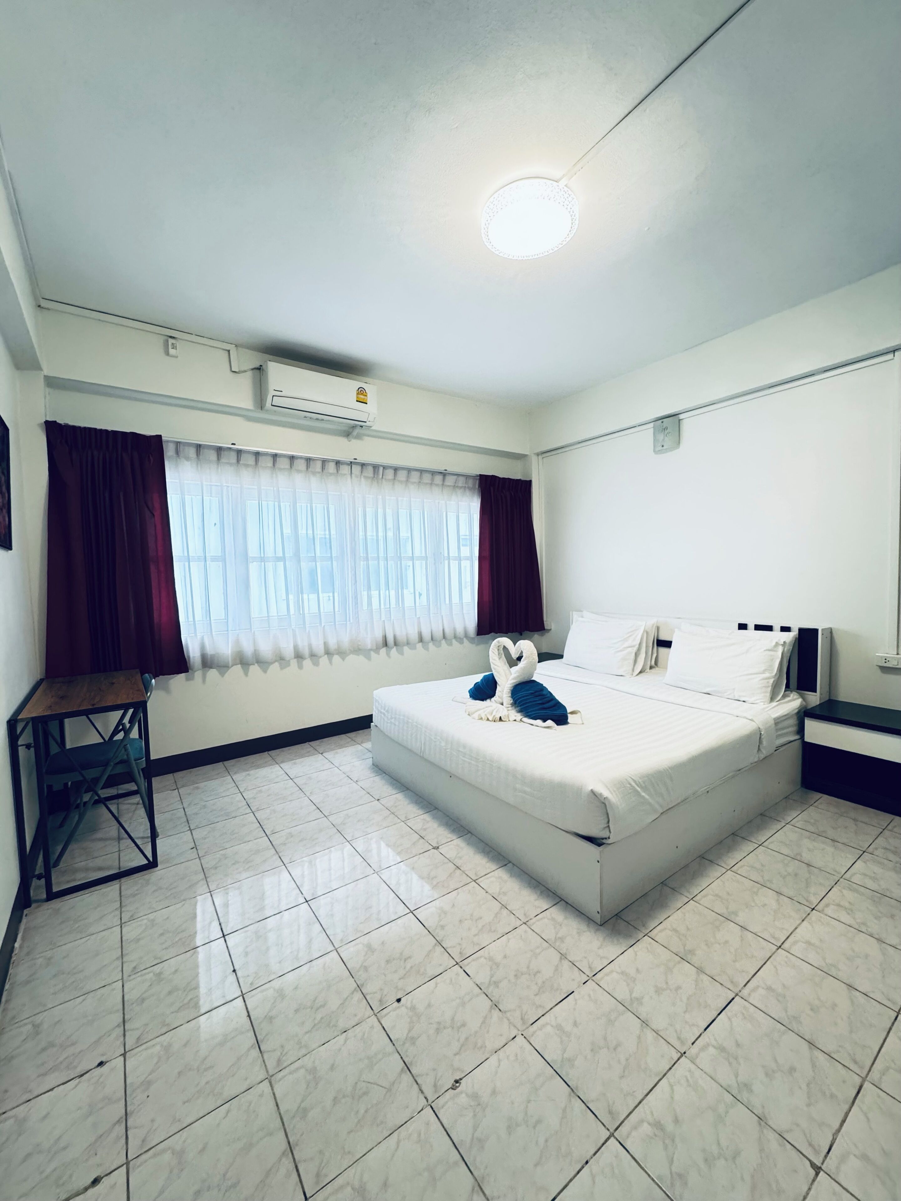 Une chambre d’hôtel comprenant un lit, un bureau, une chaise, une fenêtre avec des rideaux, un appareil de climatisation et un sol carrelé.
