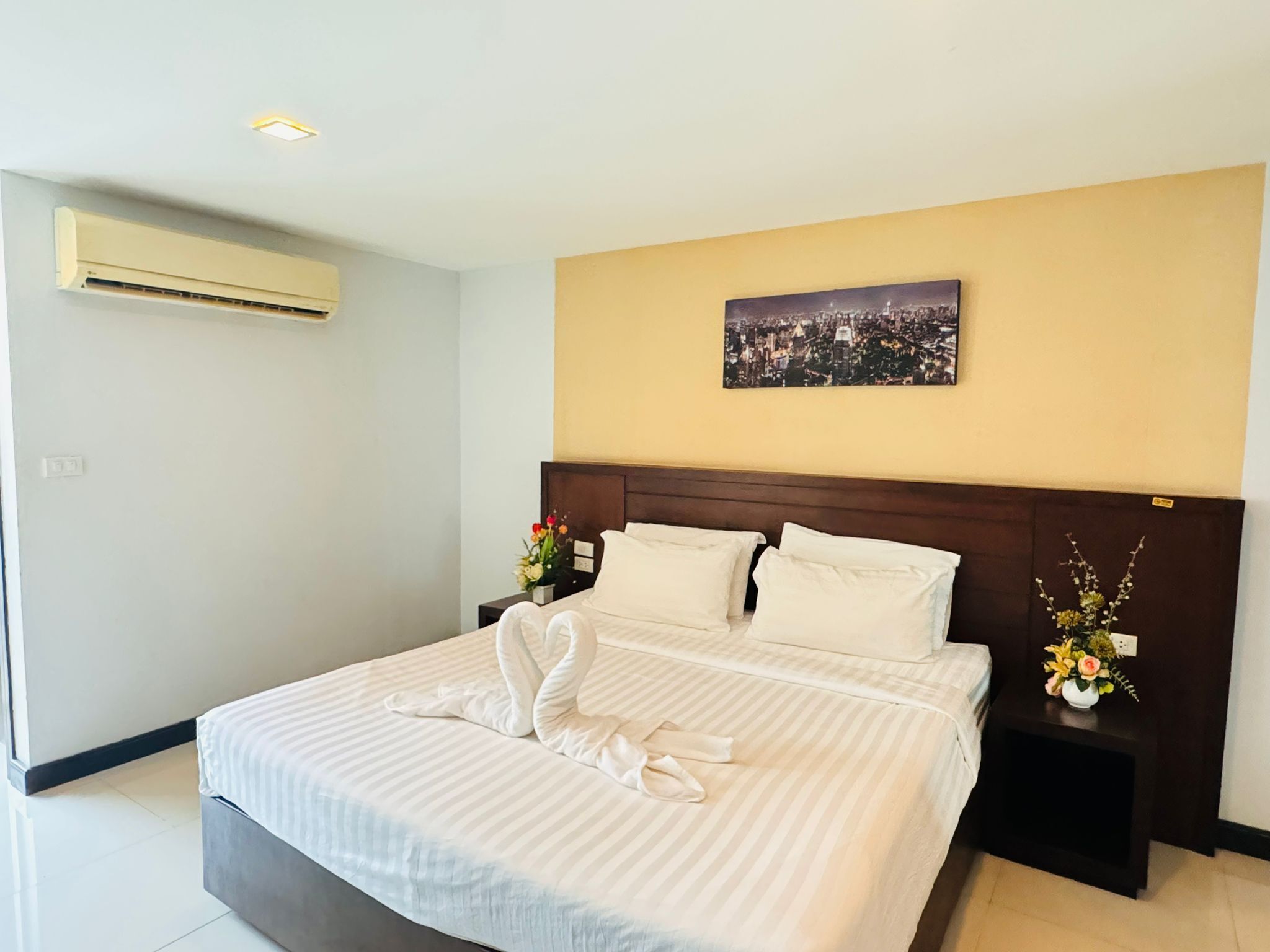 Ein Hotelzimmer mit einem großen Bett, einem Kopfteil mit Stadtansicht, einem Nachttisch mit Blumenvase und einer Klimaanlage an der Wand.
