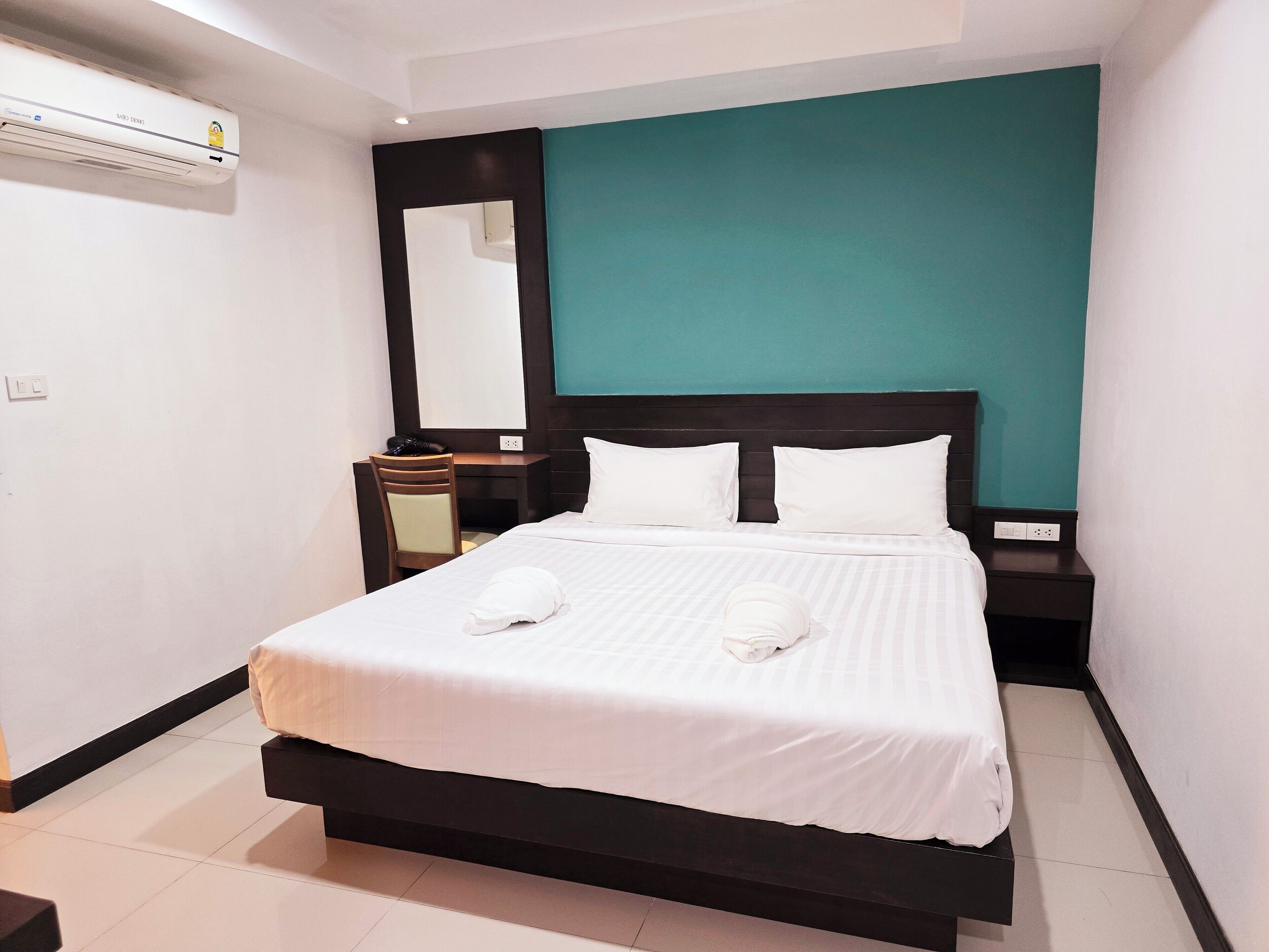 A neatly made bed with white linens and pillows, a desk with a chair, a mirror, and a teal accent wall.