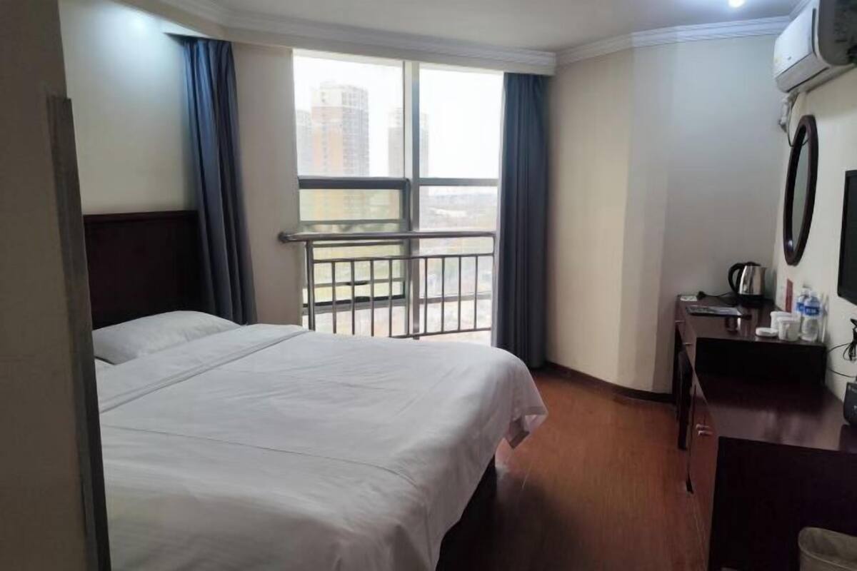 A hotel room with a bed, a desk with a chair, a small table, a window with curtains, and a view of buildings outside.