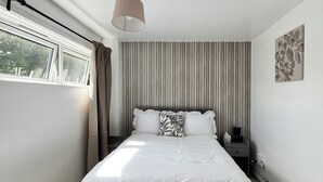 Een slaapkamer met een groot bed, een nachtkastje, een lamp, een raam met gordijnen en een aan de muur hangend kunstwerk.