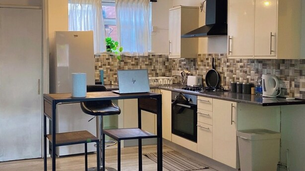 Een compacte keuken met een gasfornuis, een kleine eettafel met twee stoelen, een koelkast en een prullenbak.