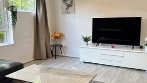 Een moderne woonkamer met een leren bank, een glazen salontafel, een flatscreen televisie op een witte kast, een grote klok aan de muur en een raam met gordijnen.