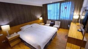 Habitación de hotel con una cama grande, dos mesitas de noche, un escritorio con silla, una mesita pequeña y una ventana con cortinas.