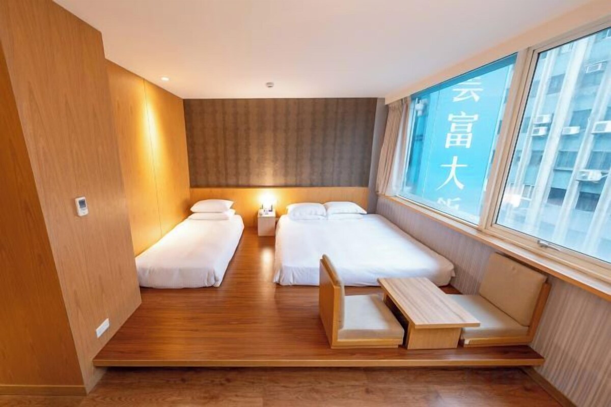 A hotel room with two beds, a wooden floor, a window with a view, and a bedside table.