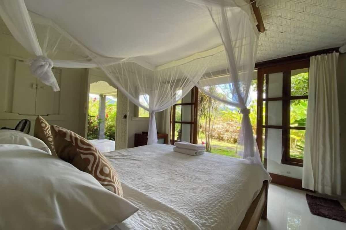 Een slaapkamer met een bed, wit beddengoed, een hemelbed en grote ramen met uitzicht op groen.