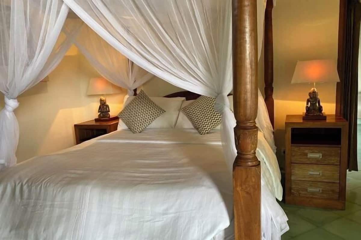 Een bed met hemelbed en houten hoofdeinde, twee nachtkastjes met lampen, gestreepte kussens en een nachtkastje met lades.