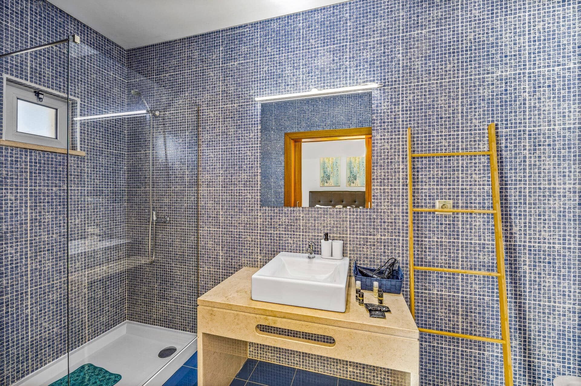 A bathroom with a tiled wall, a sink, a mirror, and a shower.