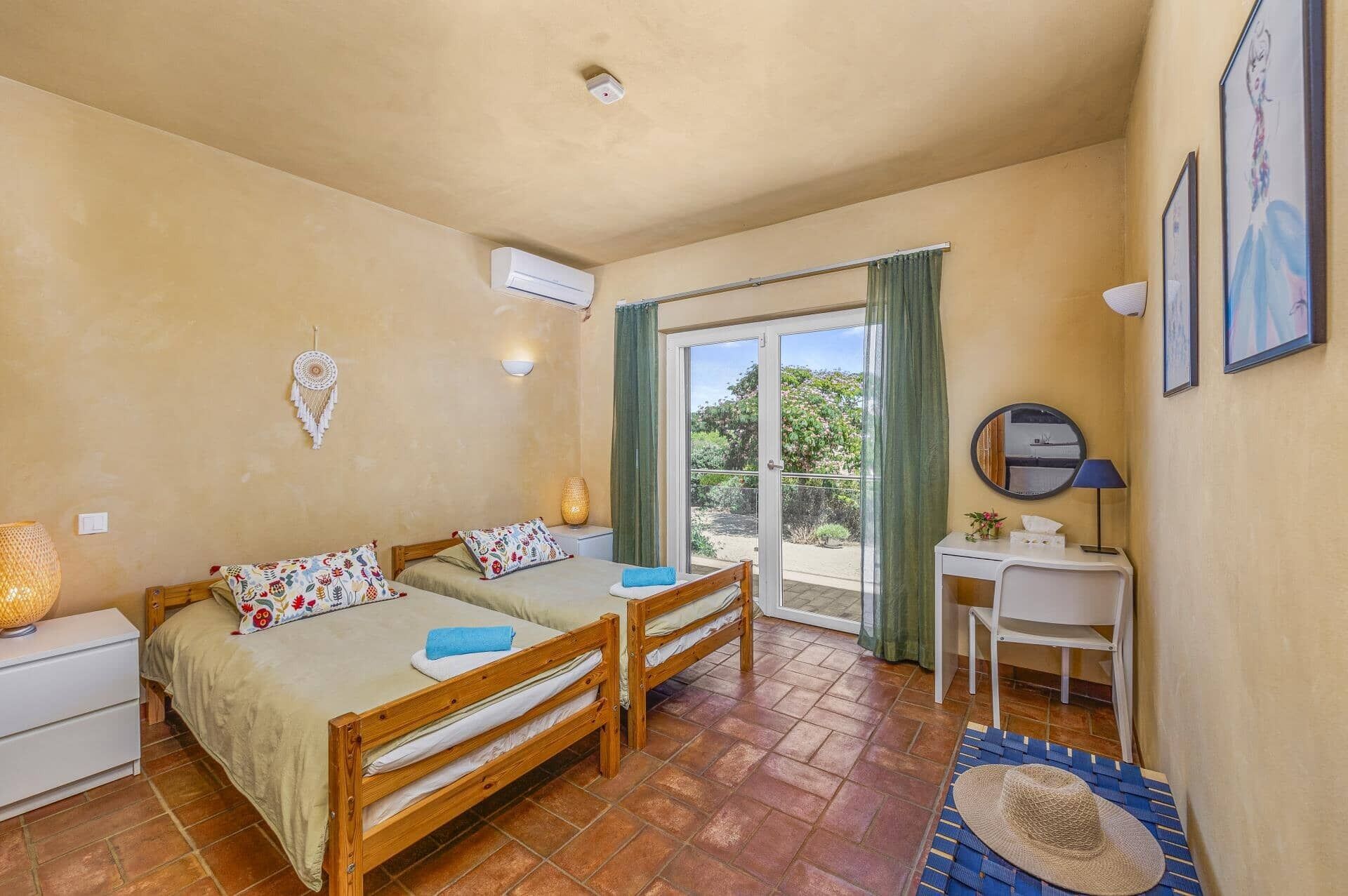 Camera da letto con due letti in legno, una scrivania, una sedia, una finestra con tende e vista su spazi verdi.