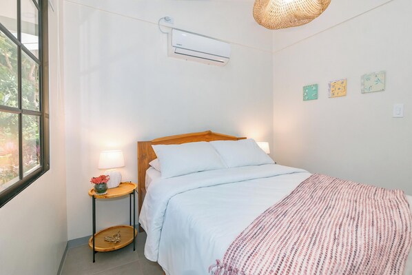 A bedroom with a bed, bedside table, lamp, air conditioning unit, and artwork on the wall.
