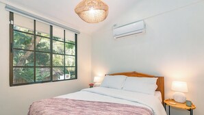 A neatly made bed with a patterned blanket, a bedside table with a lamp, and a window with a view of greenery.