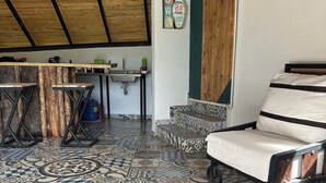 Ein Zimmer mit Fliesenboden in verschiedenen Mustern, einer Holzzielung, einem Bett mit gestreiftem Kissen, einem kleinen Tisch mit Waschbecken und einem an der Wand befestigten Schild.