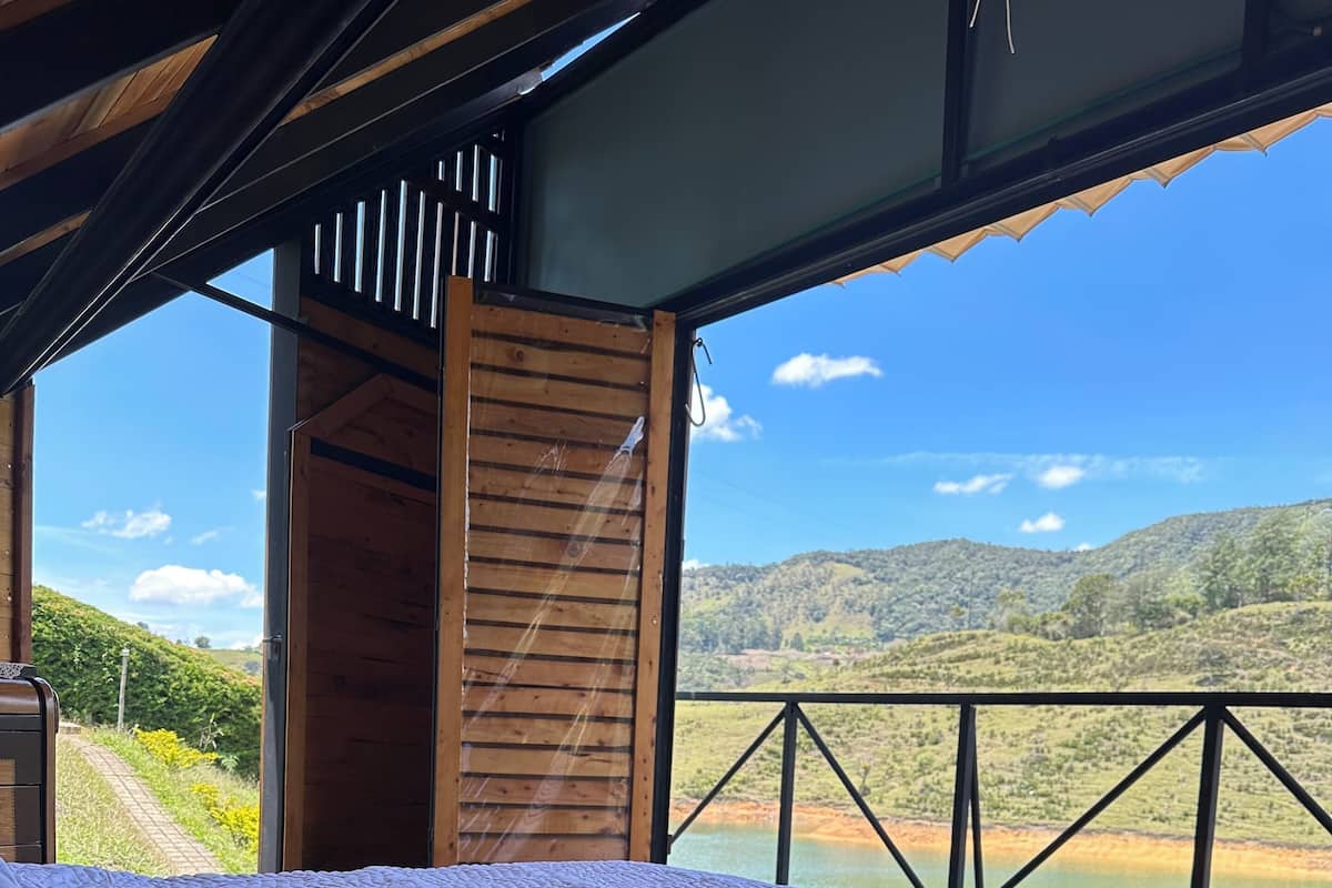 Una cama con colcha blanca en una habitación con piso de madera y un amplio ventanal con vistas a un paisaje pintoresco.
