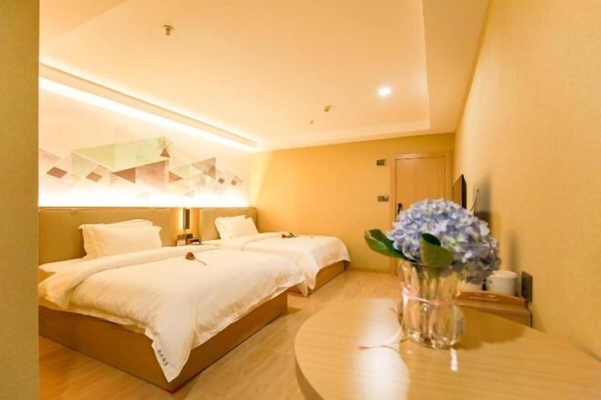 A hotel room with two beds, a bedside table with a vase of flowers, and a wall-mounted lamp.