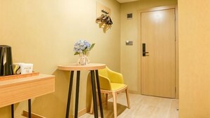 Quarto de hotel com escrivaninha de madeira, uma cadeira, uma mesinha com um vaso de flores e uma porta.