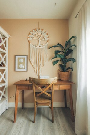 A room with a wooden desk, a chair, a macrame wall hanging, a framed picture, and a potted plant.