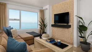 Una sala de estar moderna con un ventanal grande, un televisor de pantalla plana montado en una pared con diseño de ladrillo, un sofá con cojines, una mesa de centro con libros y una vela, y una planta en maceta.