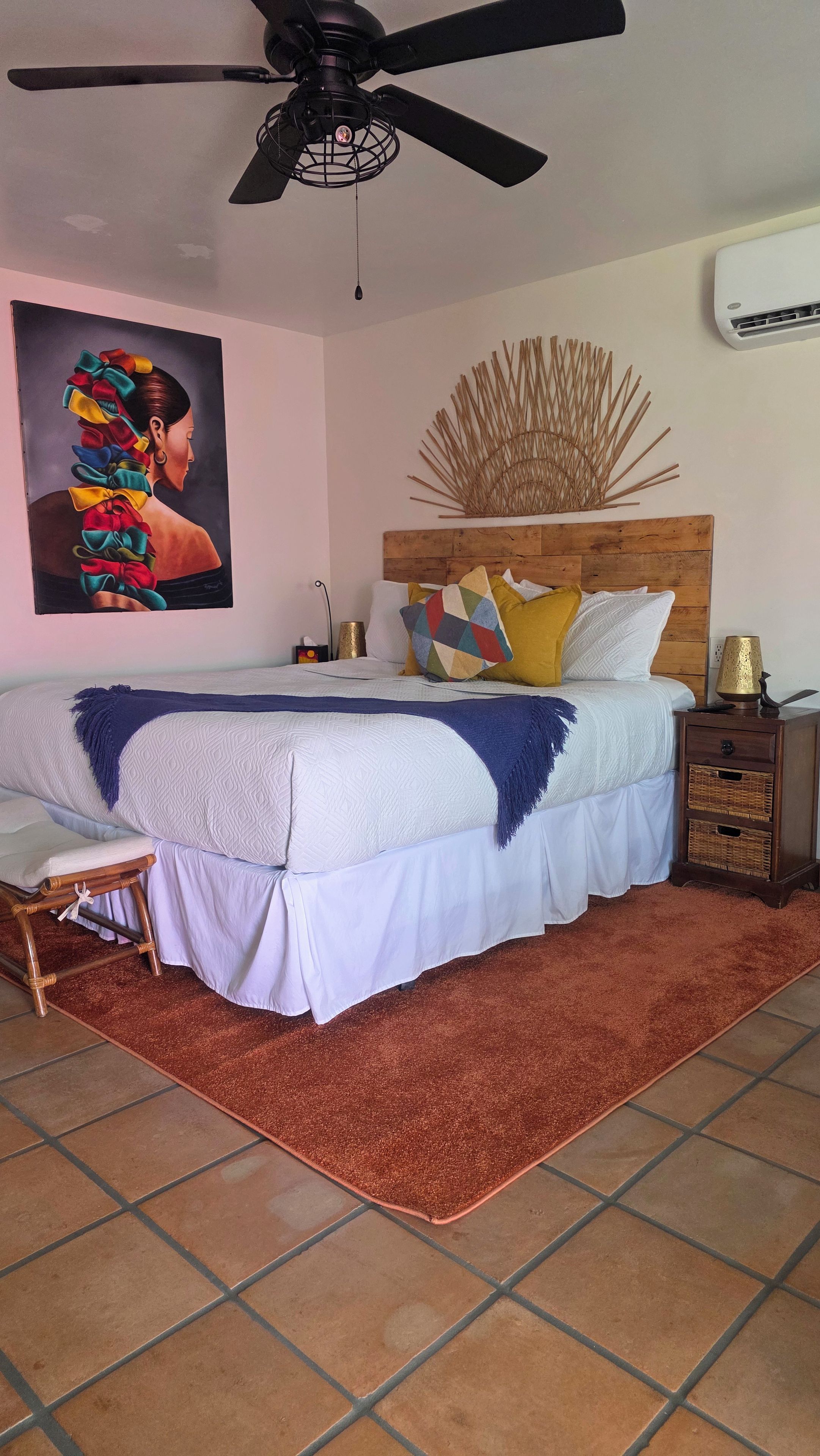 A bedroom with a bed, a ceiling fan, a wall-mounted air conditioner, a decorative wall piece, and a bedside lamp.