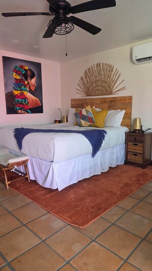 A bedroom with a bed, a ceiling fan, a wall-mounted air conditioner, a decorative wall piece, and a bedside lamp.