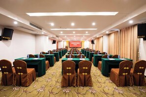 Une salle de conférence avec des rangées de tables et de chaises, une scène avec un écran et un sol recouvert de moquette.