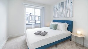 A bedroom with a large bed, a nightstand, a window with a view of buildings, and a wall-mounted artwork. - NEW Home, Double Car Garage & Close to Airport (Calgary)