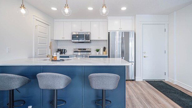 A modern kitchen with white cabinets, a stainless steel refrigerator, a microwave, and a large island with bar stools. - NEW Home, Double Car Garage & Close to Airport (Calgary)