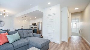 A modern living room with a grey sectional sofa, a kitchen in the background, and a hallway leading to another room. - NEW Home, Double Car Garage & Close to Airport (Calgary)