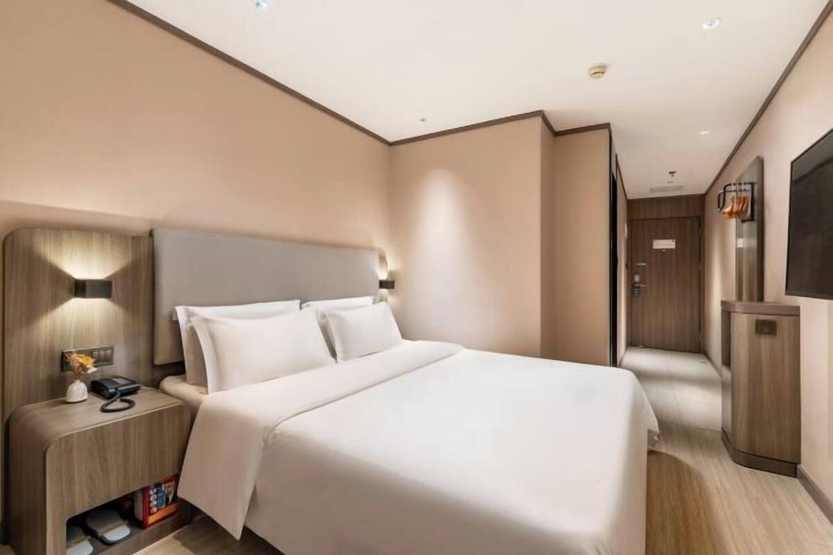 A modern hotel room with a large bed, a bedside table with a lamp, and a flat-screen TV mounted on the wall.
