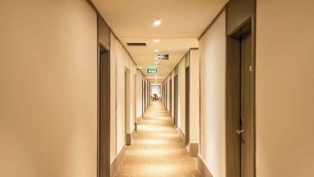 Un long couloir étroit, typique des hôtels, avec des portes des deux côtés, un sol recouvert de moquette et des panneaux indicateurs de sortie au plafond.