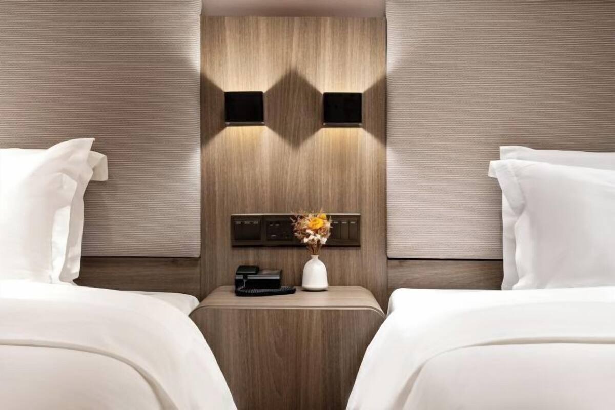 A hotel room with two beds, a nightstand with a phone and a vase of flowers, and a wall-mounted control panel.